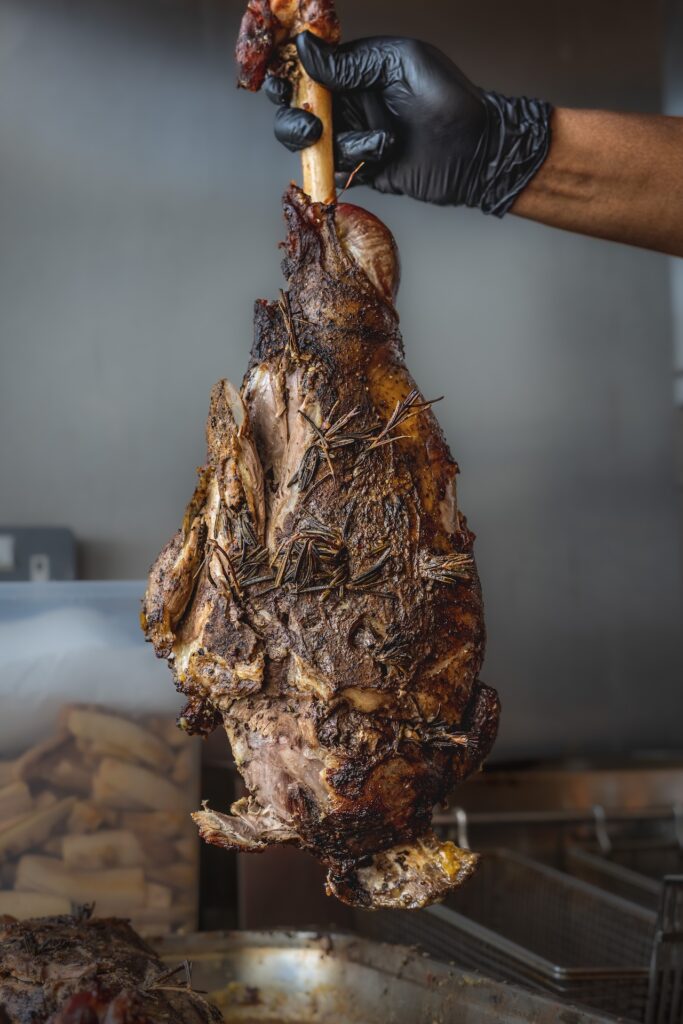 A hand holding a large, slow-cooked, bone-in cut of meat, seasoned with herbs, in a kitchen setting.