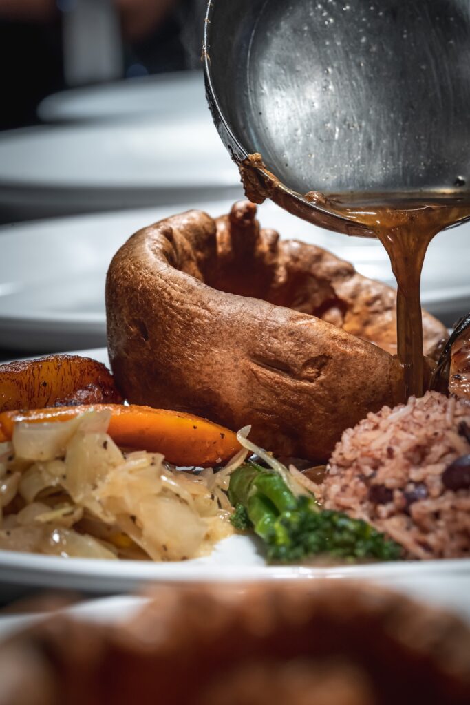 Gravy being poured from a ladle over a golden Yorkshire pudding, with sides of vegetables and rice on the plate.