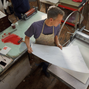 An artist holding a large sheet of paper in a studio, surrounded by printmaking equipment and red ink.