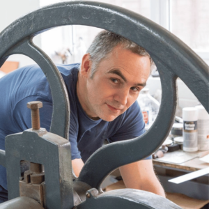 A printmaker leaning through a large printing press wheel in a workshop filled with tools and materials.