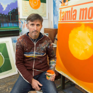 A painter in a splattered sweater holding a jar of orange paint, seated in front of vibrant artwork.