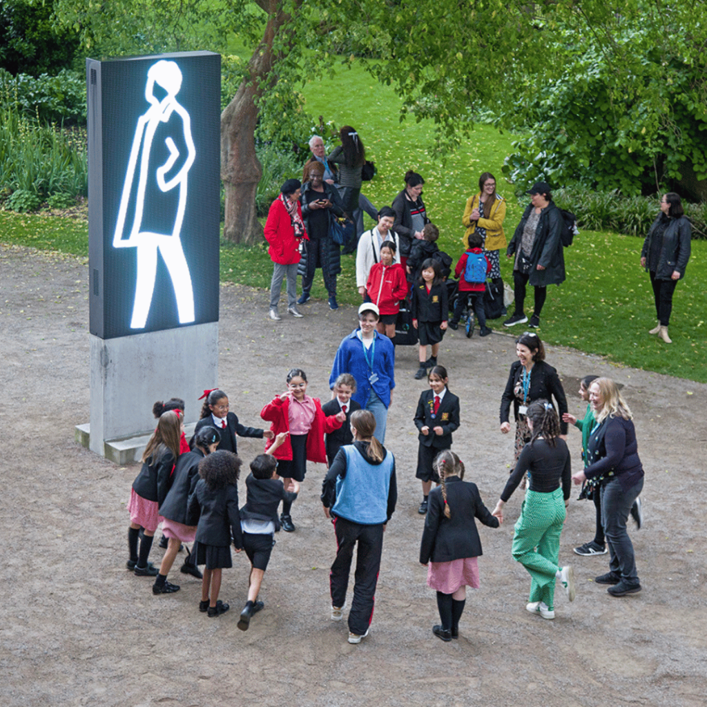 Children and adults gather outdoors, joyfully dancing in a ring near Julian Opie's illuminated artwork.