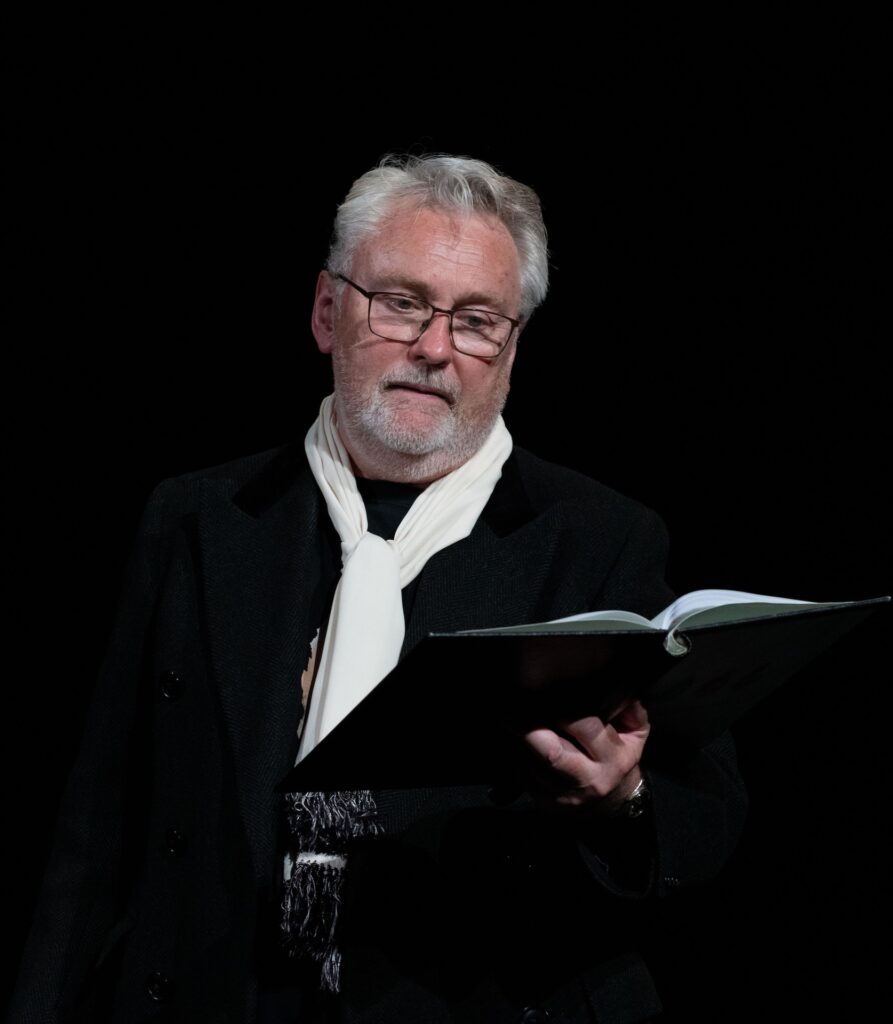 A bearded actor in a black coat and white scarf solemnly recites from a book in a dimly lit theatre.