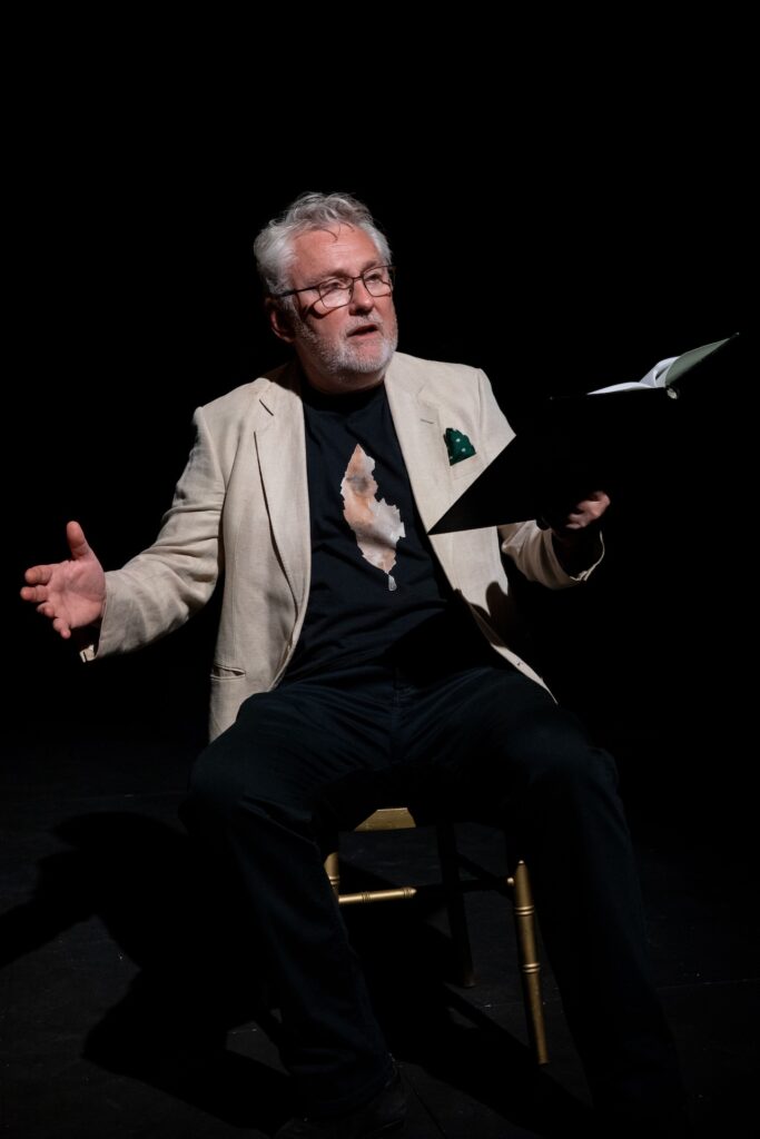 A male actor in a tropical shirt looks upward, holding a script, illuminated against a dark stage.